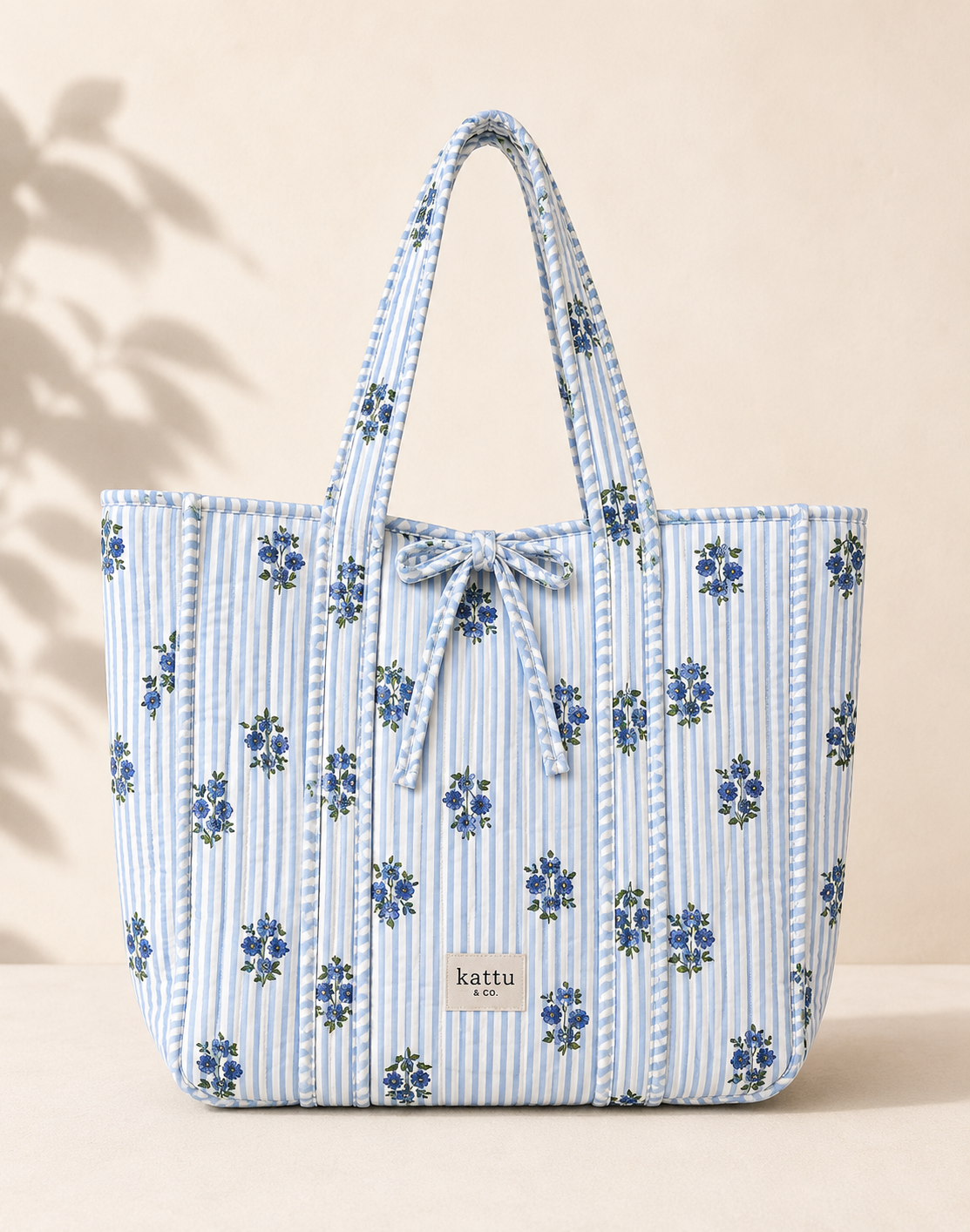 A quilted tote bag with a blue and white striped fabric body, decorated with small blue floral motifs, featuring two top handles and a drawstring bow at the top center. A small rectangular brand label reading 'kattu' is visible on the front.